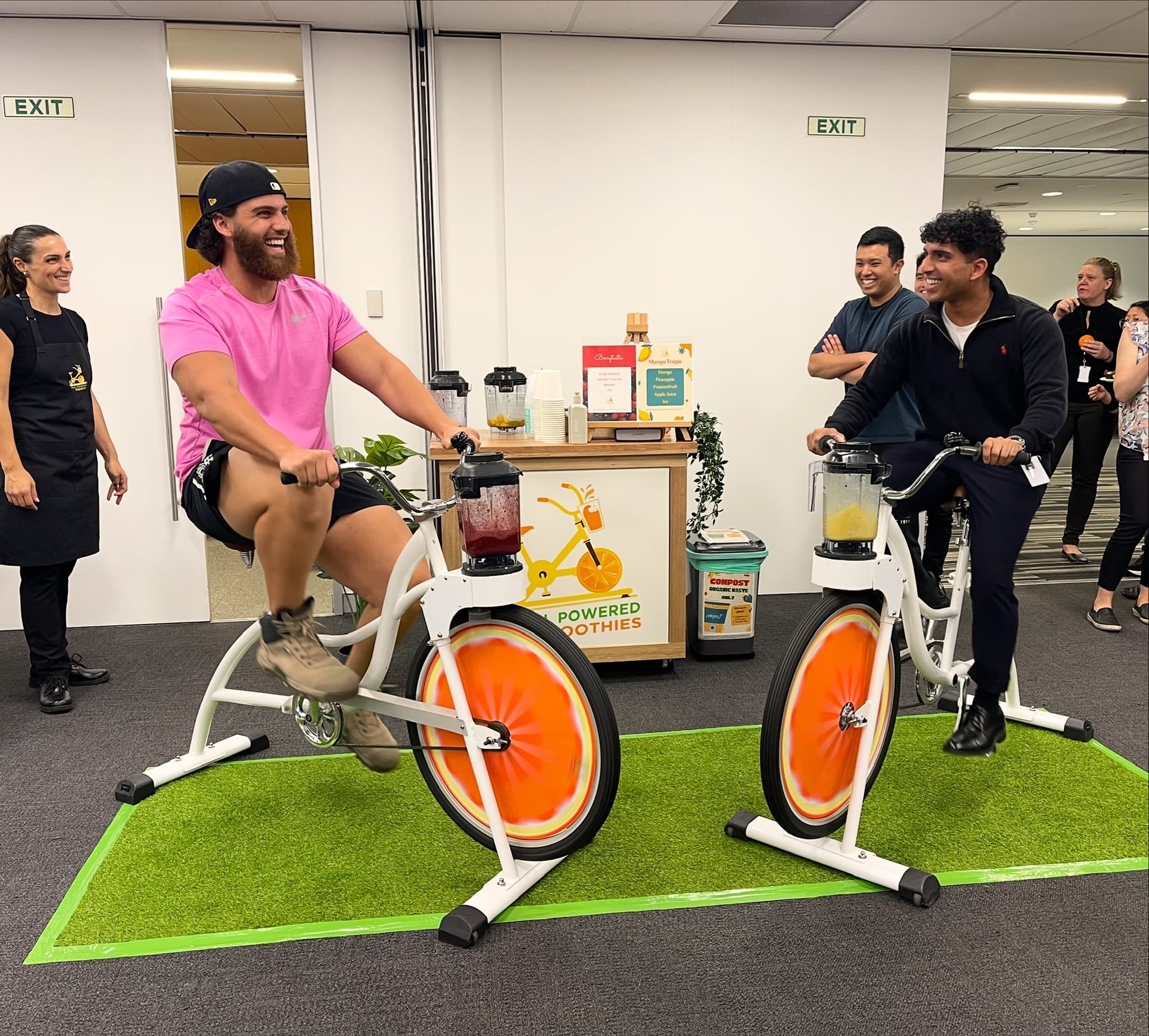 Two men are sitting on stationary bikes in a modern office hallway, pedalling vigorously to power blenders attached to the handlebars. The man on the left has a magnificent beard and wears a pink shirt and black cap, laughing as he pedals, while the man on the right wears a black pullover. Others watch with amusement in the background.