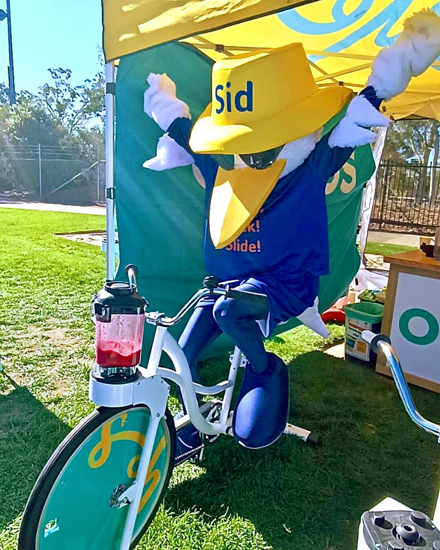 A mascot named "Sid"—a large yellow bird wearing a sun hat (labelled "Sid") and sunglasses—enthusiastically pedals a white smoothie bike under a yellow tent. A blender filled with a red berry smoothie is mounted to the front of the bike.