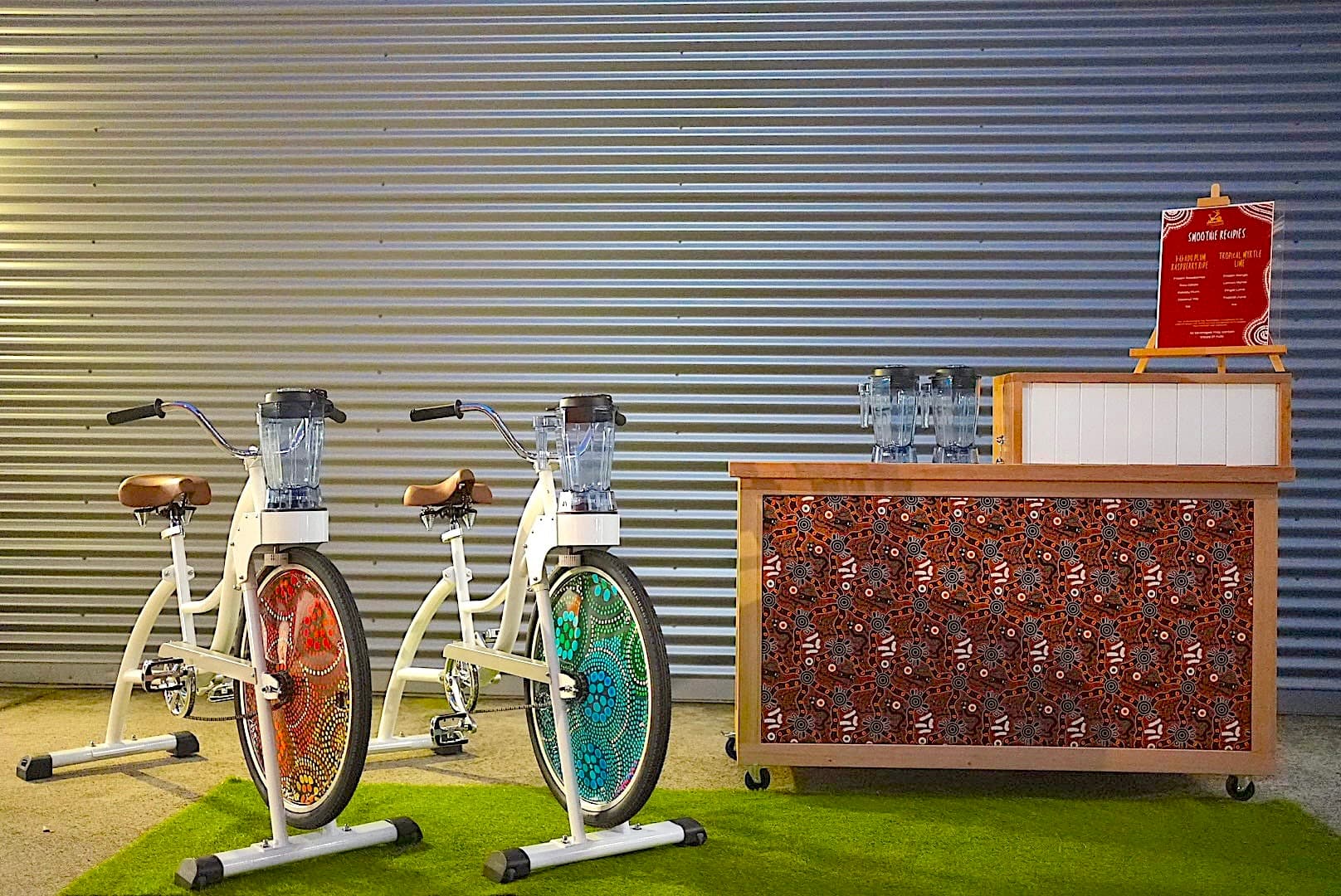 Two stationary bikes with colorful wheel designs are equipped with blenders. They are next to a counter with Aboriginal art, on a green rug.