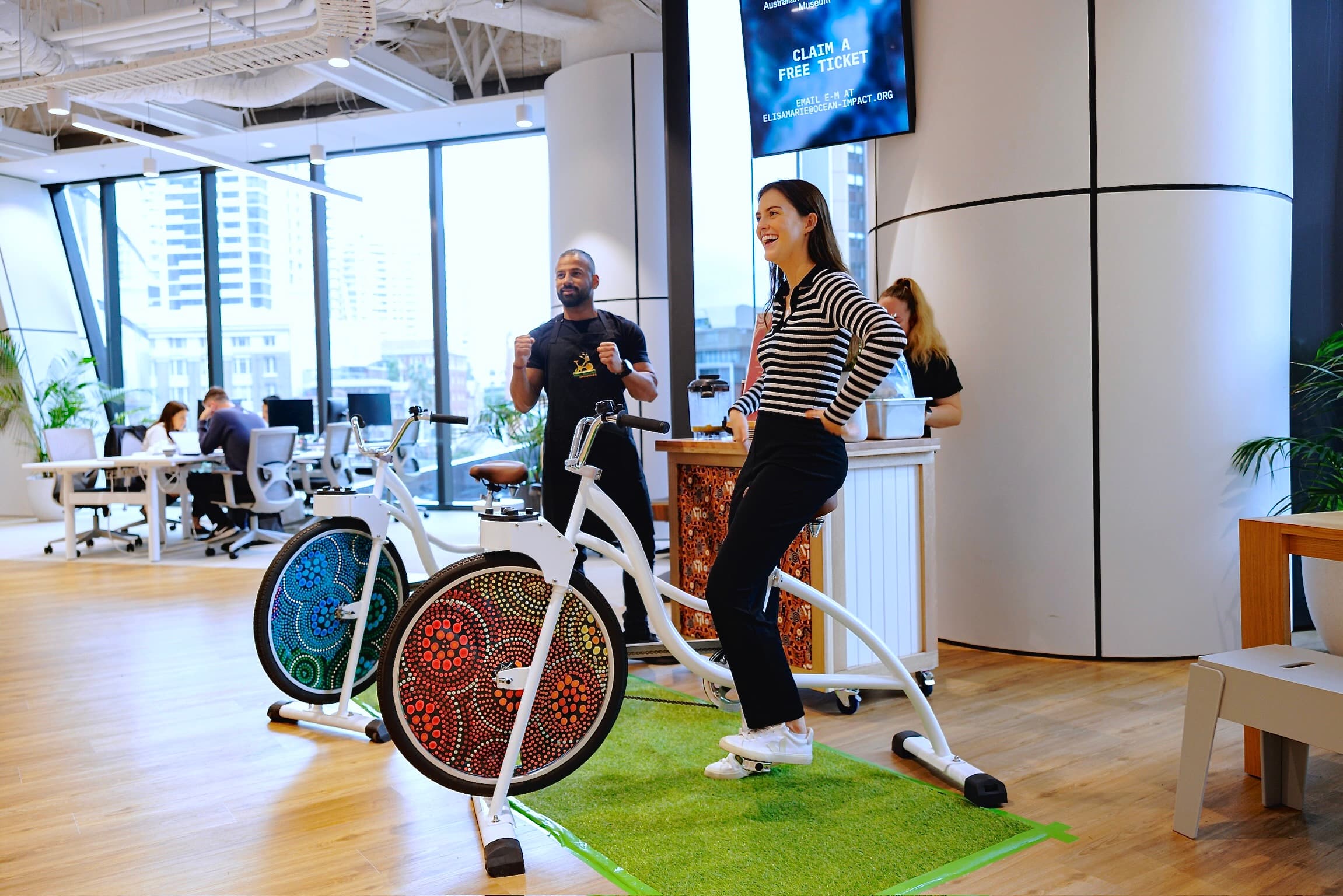 A woman in a striped shirt laughs while pedalling a white blender bike in a bright, modern office or event space with large windows and curved white pillars. Behind her, a staff member in a black apron cheers her on. The bikes feature colorful, indigenous-style dot art on the front wheel guards.