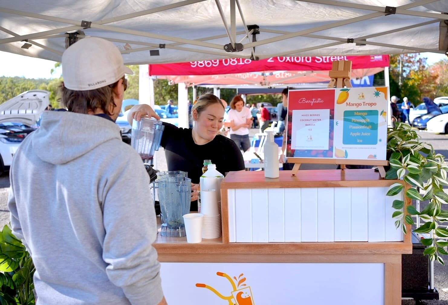 A close-up shot of a staff member behind a wooden mobile bar, pouring a freshly blended smoothie from a blender jug into a cup for a customer. A menu on the counter lists flavors like "Berrytastic" and "Mango Tropo". The scene takes place outdoors under a white pop-up gazebo at a busy event.