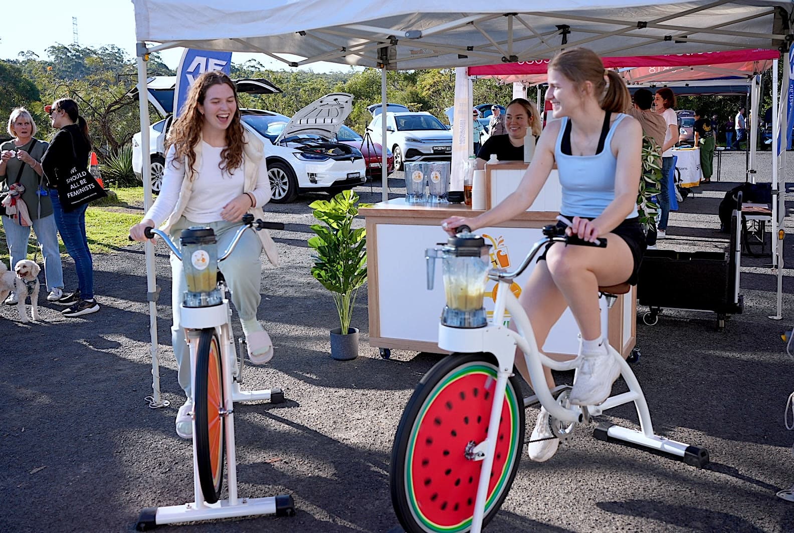 Two young women are laughing as they pedal stationary blender bikes at an outdoor event. The bikes are white with colorful fruit-themed wheel covers (orange and watermelon). A mobile smoothie bar stands behind them under a white canopy, with several cars and event-goers visible in the background.