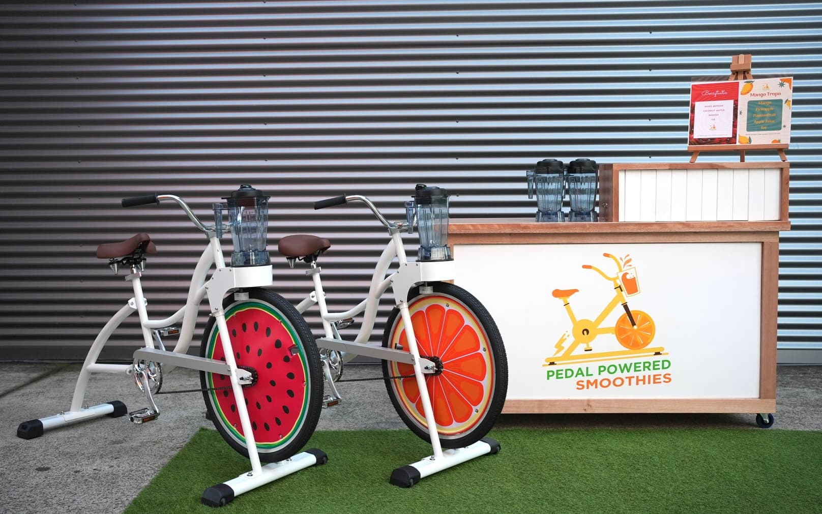 A clean, wide shot of two stationary blender bikes positioned in front of a wooden mobile bar. The bar features a logo that says "PEDAL POWERED SMOOTHIES." One bike has a watermelon-patterned wheel, and the other has an orange-slice pattern. They are set up on a patch of artificial grass against a grey corrugated metal wall.