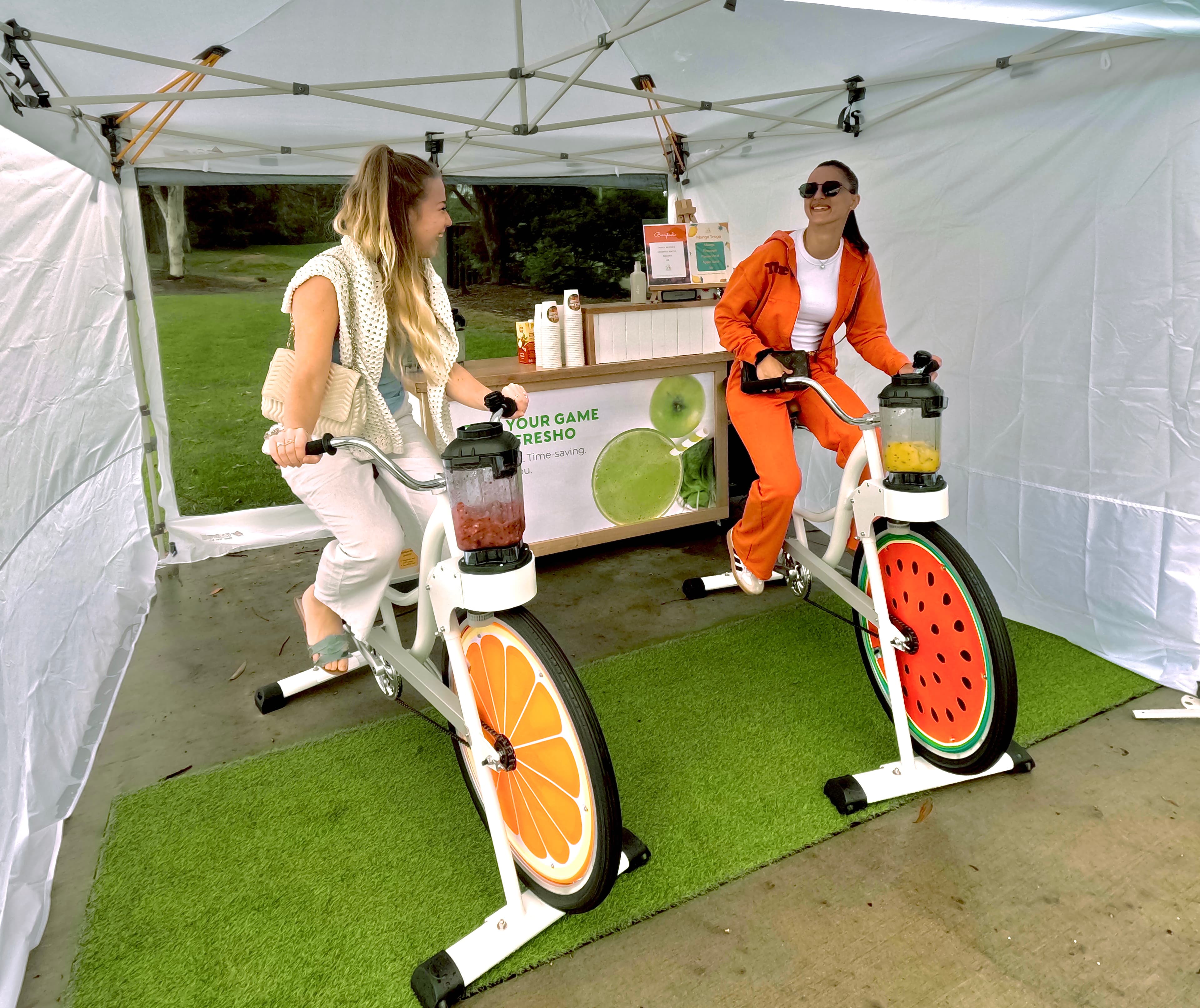 Two women laugh while using smoothie bikes inside a white gazebo at an outdoor event. The bike wheels are decorated with vibrant orange and watermelon fruit slices. A wooden bar in the background features "Fuel Your Game" signage.