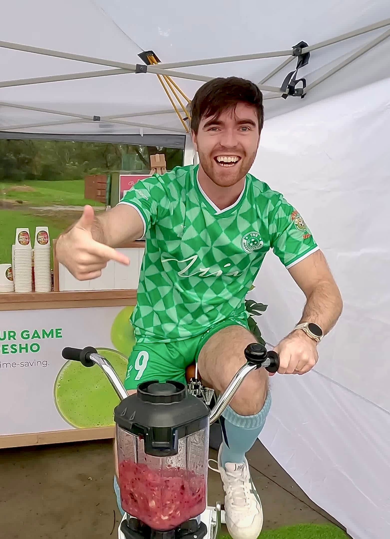 A man in a bright green soccer jersey gives a thumbs-up and points toward the camera while pedalling a smoothie bike. A blender full of red fruit is being mixed by his pedalling action.