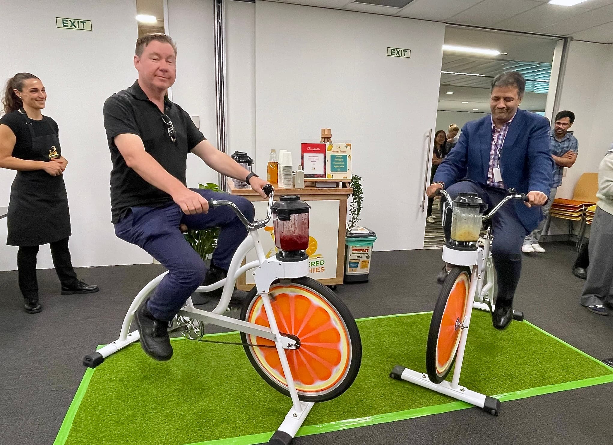 Two men in professional attire are pedalling white stationary bikes in an office setting to power blenders attached to the handlebars. The man on the left wears a black polo shirt, and the man on the right wears a blue blazer. A staff member in a black apron stands to the left, watching with a smile.