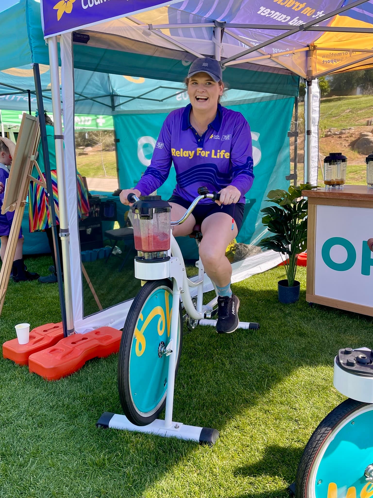 A person in a "Relay For Life" shirt joyfully pedals a stationary bike blender under a tent at an outdoor event. Bright, sunny atmosphere.