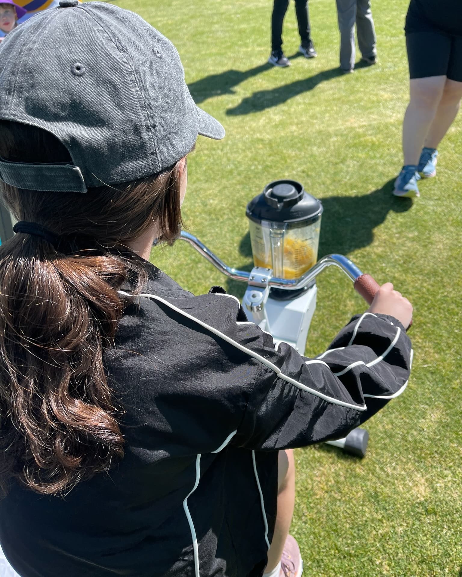 An over-the-shoulder view of a young girl wearing a black jacket and grey cap, riding a white bike. A blender filled with a yellow smoothie is mounted to the handlebars, powered by the pedalling motion, set outdoors on a sunny grass field.