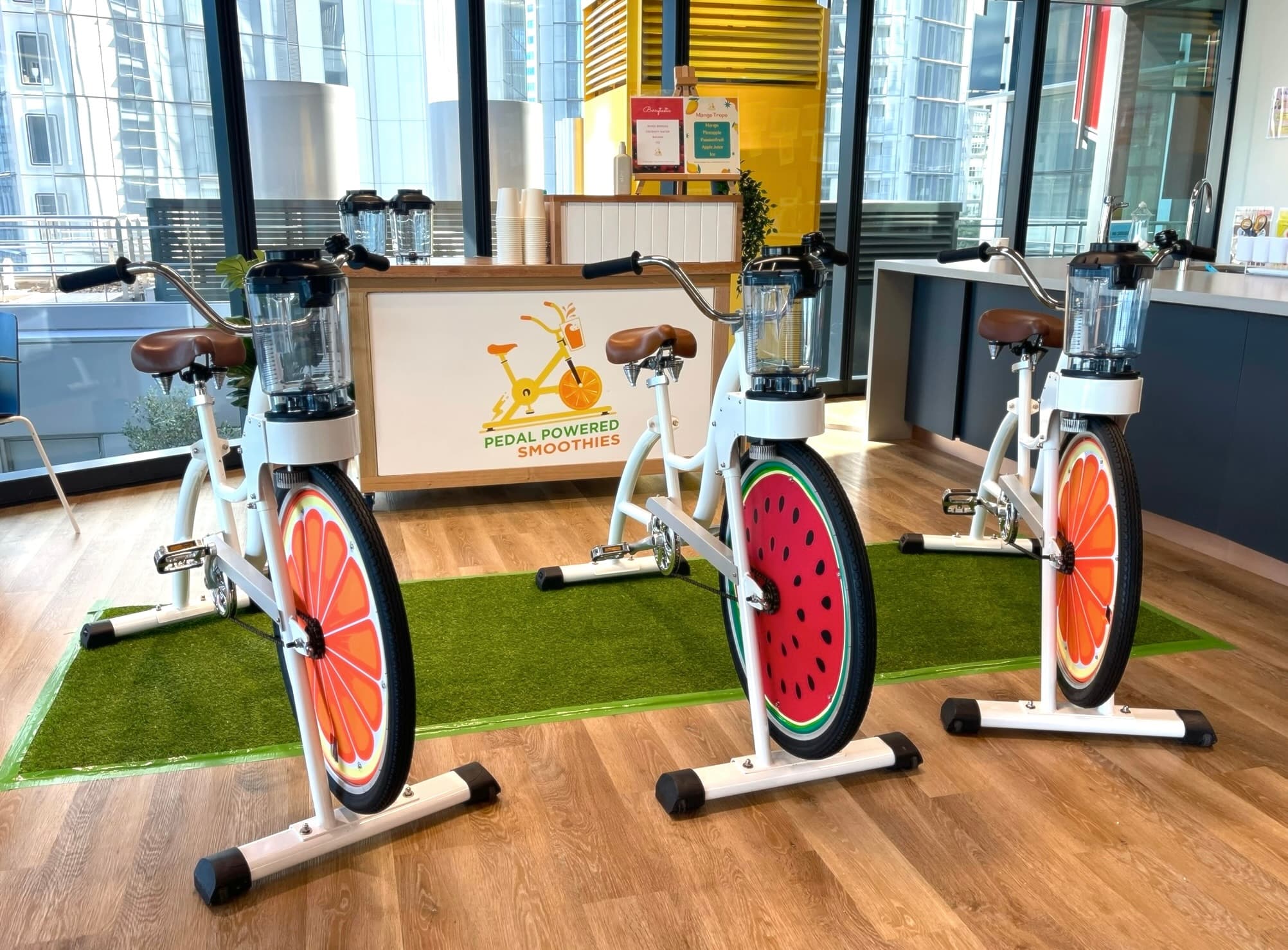 A trio of white stationary blender bikes are lined up on a green artificial turf mat inside a modern, high-rise office with floor-to-ceiling windows. The wooden service bar in the background displays the "Pedal Powered Smoothies" logo and a menu. Each bike is equipped with a blender jug and features vibrant fruit-graphic wheels.