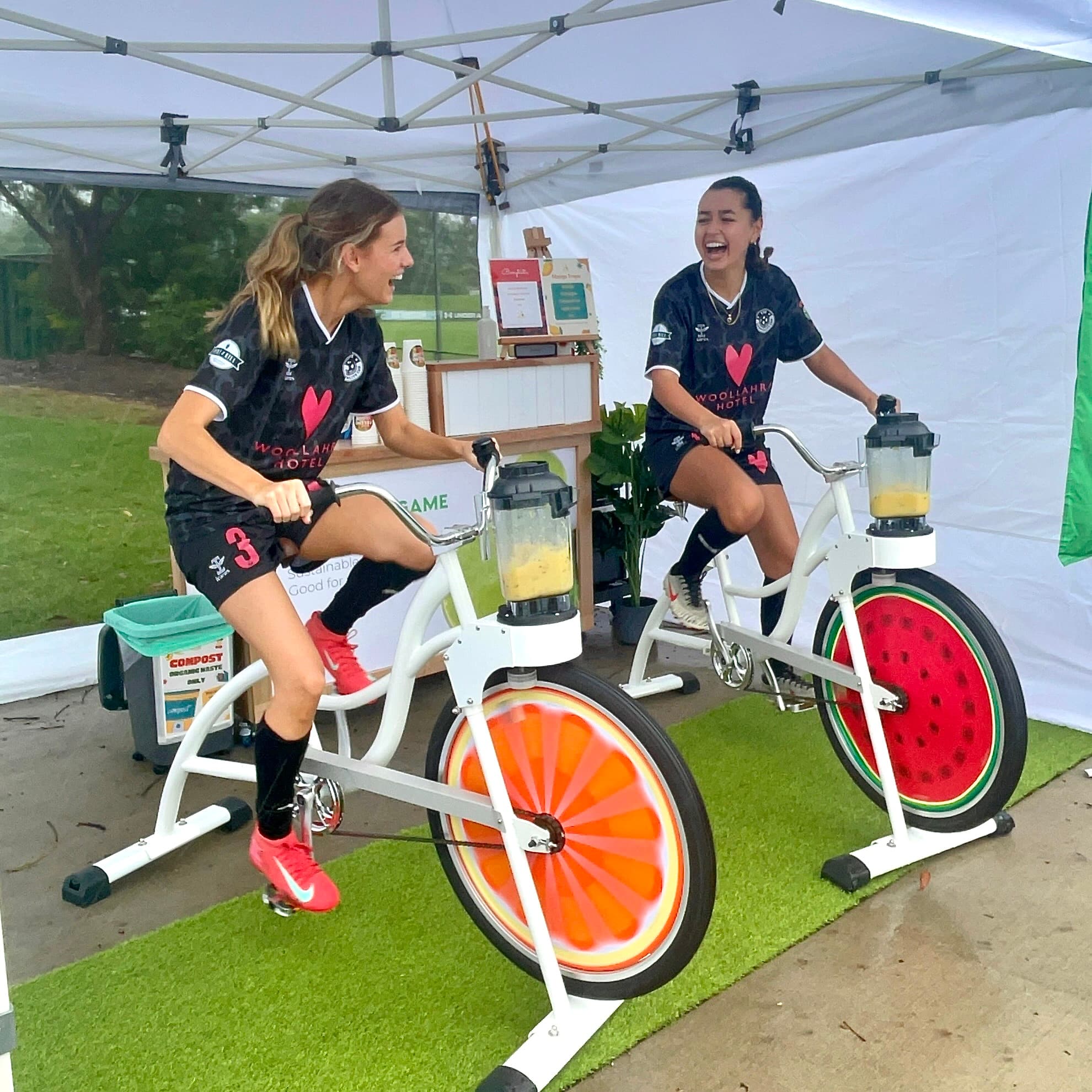 Two women in black athletic gear with large pink hearts on the front are laughing together as they pedal fruit-slice stationary bikes side-by-side under a white marquee on a wet day.