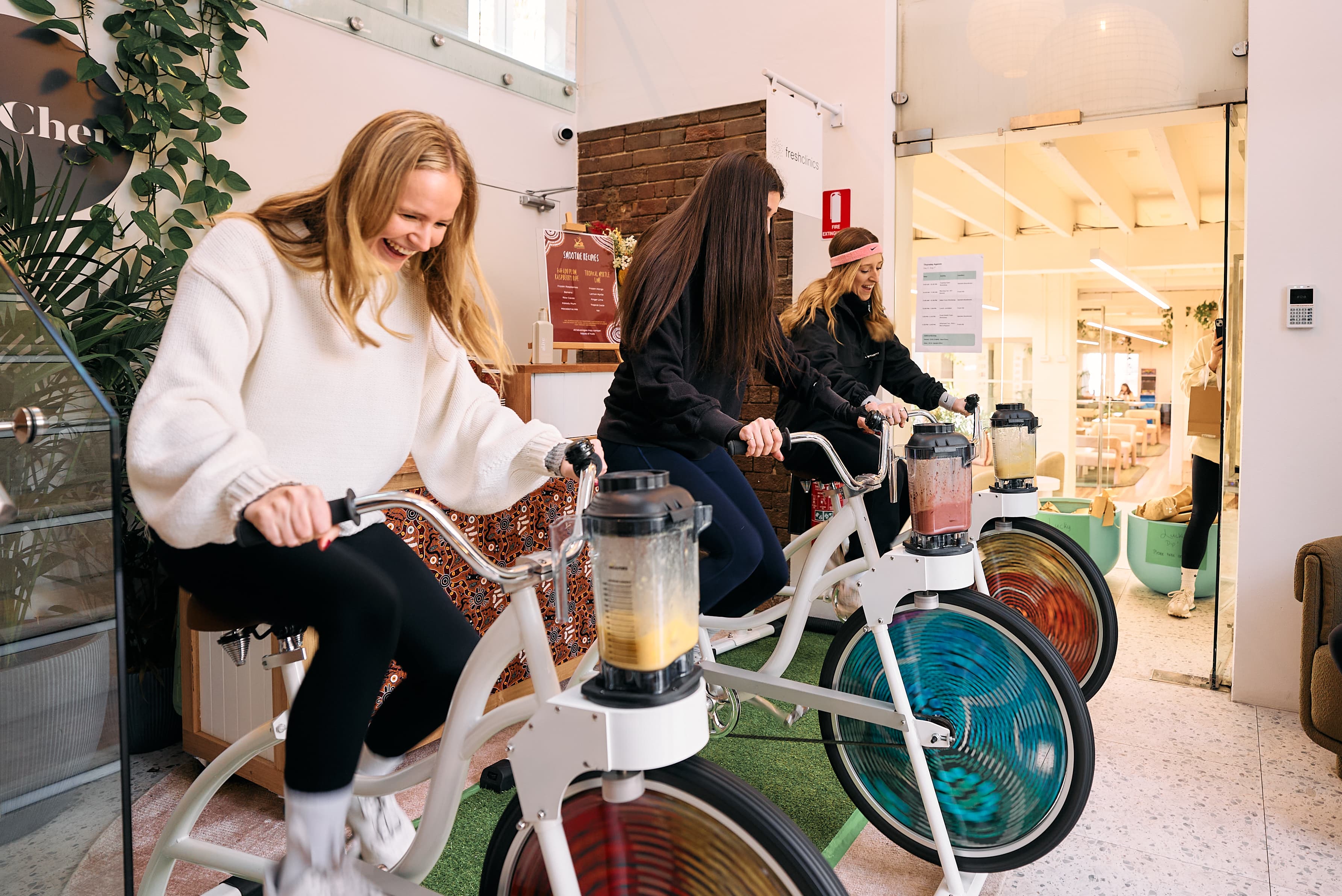 Three women are lined up on blender bikes, pedalling in unison during an office wellness event. The woman in the foreground wears a cozy white sweater and is laughing as she watches her yellow smoothie blend. The bikes are set up on a green turf mat in a bright, modern entryway with exposed brick accents.