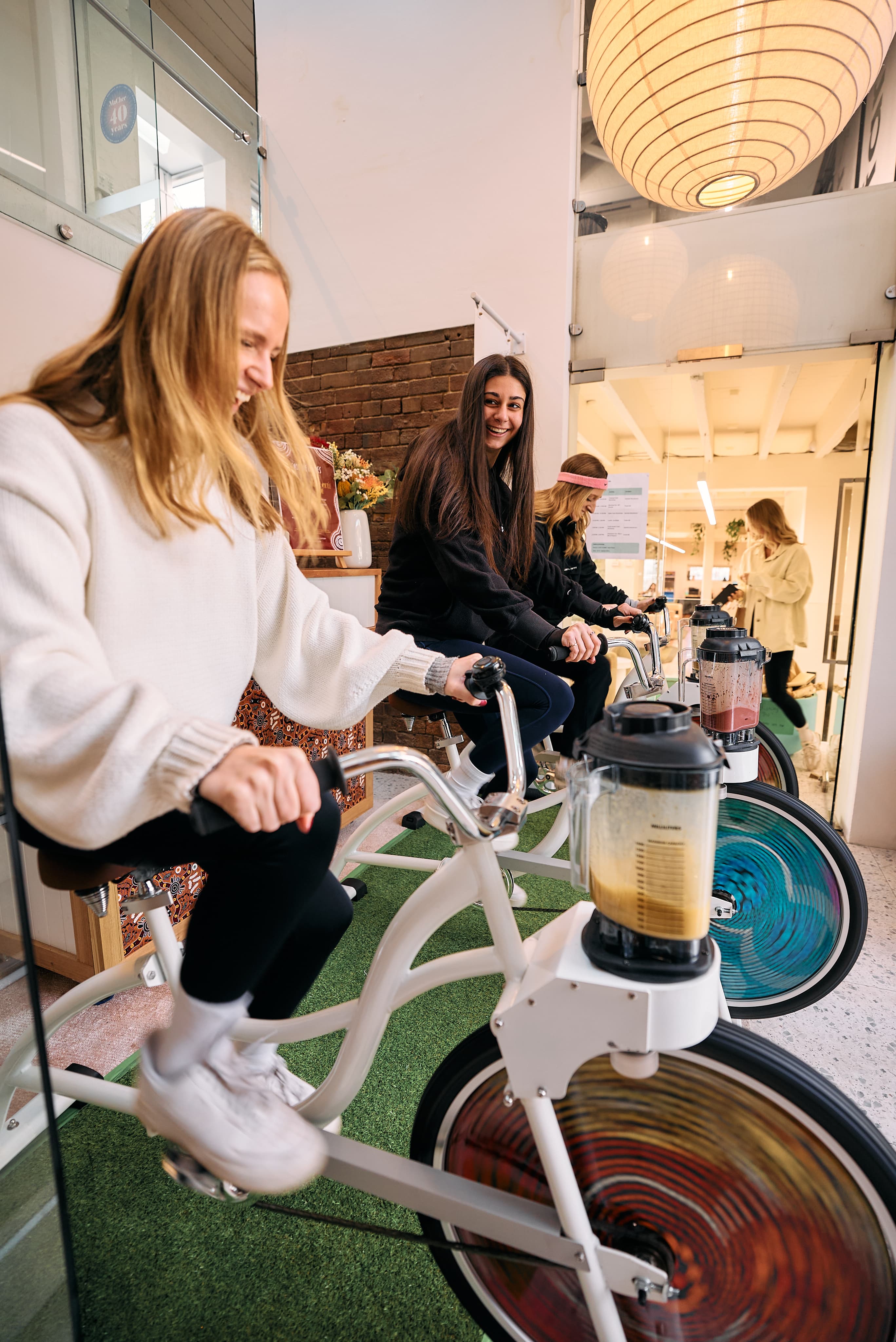 Three women smile as they pedal stationary blender bikes indoors on a strip of artificial turf. Blenders attached to the front of the white bikes spin to mix smoothies as they pedal.