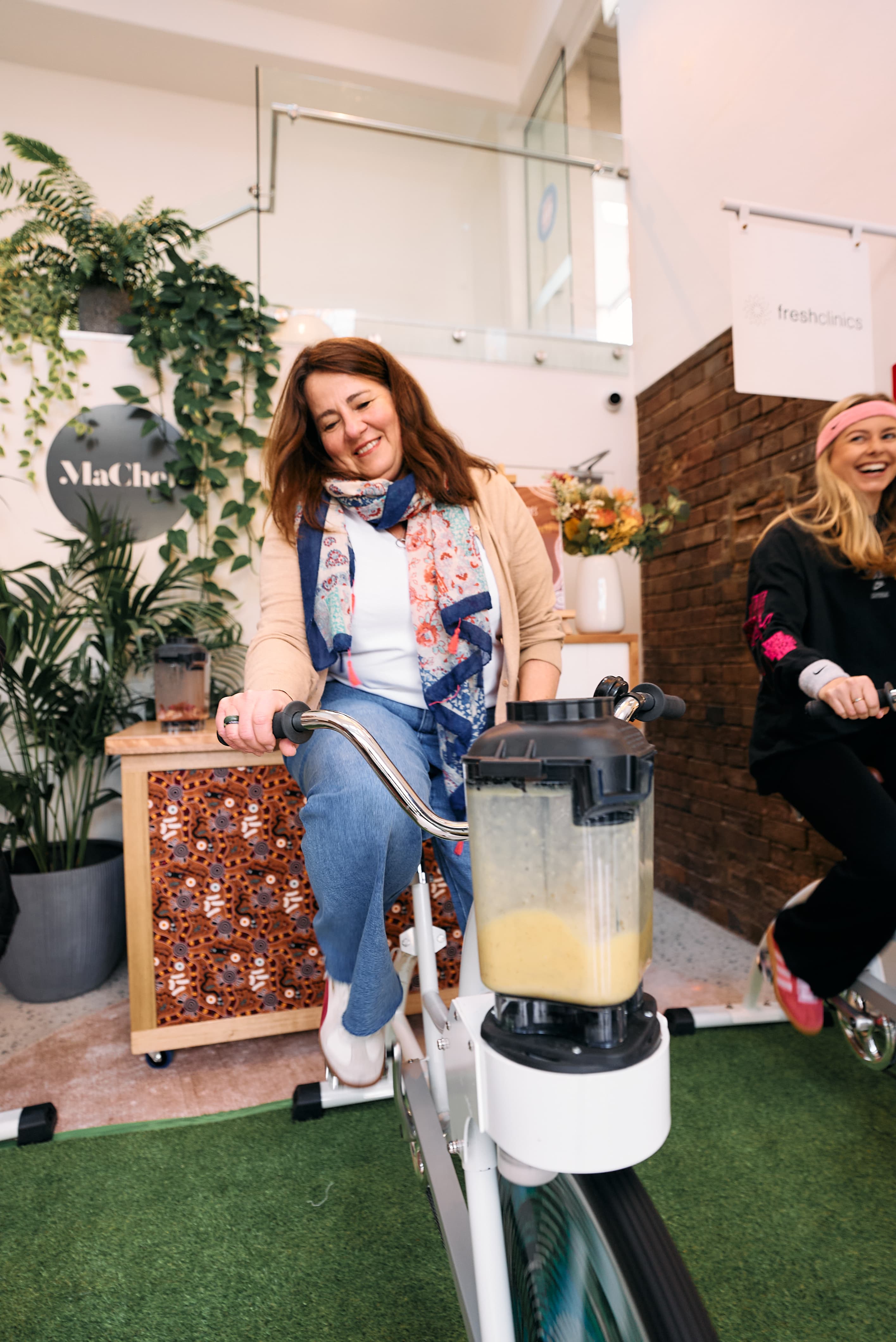Two women smiling and pedalling stationary bikes with colorful fruit designs, under a white marquee at an outdoor event. They're blending smoothies as they cycle.